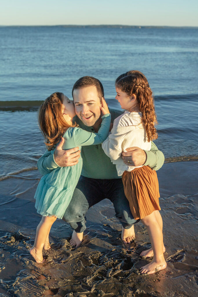 A smiling dad holds his little girls in his arms as one pulls him in to give him a kiss on the cheek. The other little girl smiles at her dad and little sister, the ocean waves lapping gently in the background.