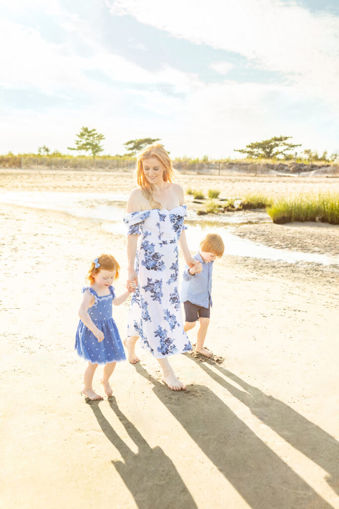 A mother wearing a white and blue off-the-shoulder dress walks along a Connecticut beach with her young daughter and son. The trio's shadows extend out in front of them as if they are chasing their shadows.