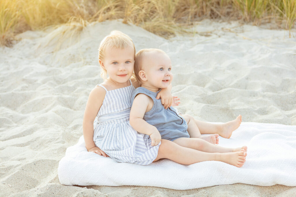 A proud big sister holds her baby brother (who is almost as big as she is!) in her lap as the pair sit on a folded white textured beach blanket, on the soft sands of Greenwich Beach.
