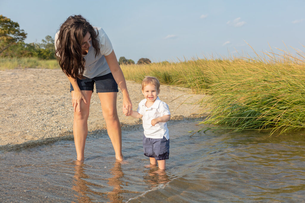 A mom thicky, shiny dark hair holds the hand of her toddler son and smiles down at him. The pair are standing in the shallow water of Greenwich Beach and the little boy smiles broadly, the hem of his shorts wet with saltwater.
