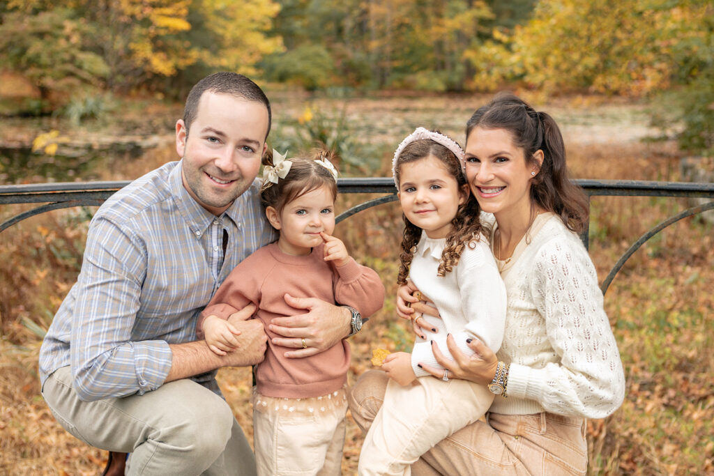 A family of four is photographed during a fall foliage family session in the fall of 2021. Mom and Dad each hold onto one of their daughters, all four of them smiling at the camera.