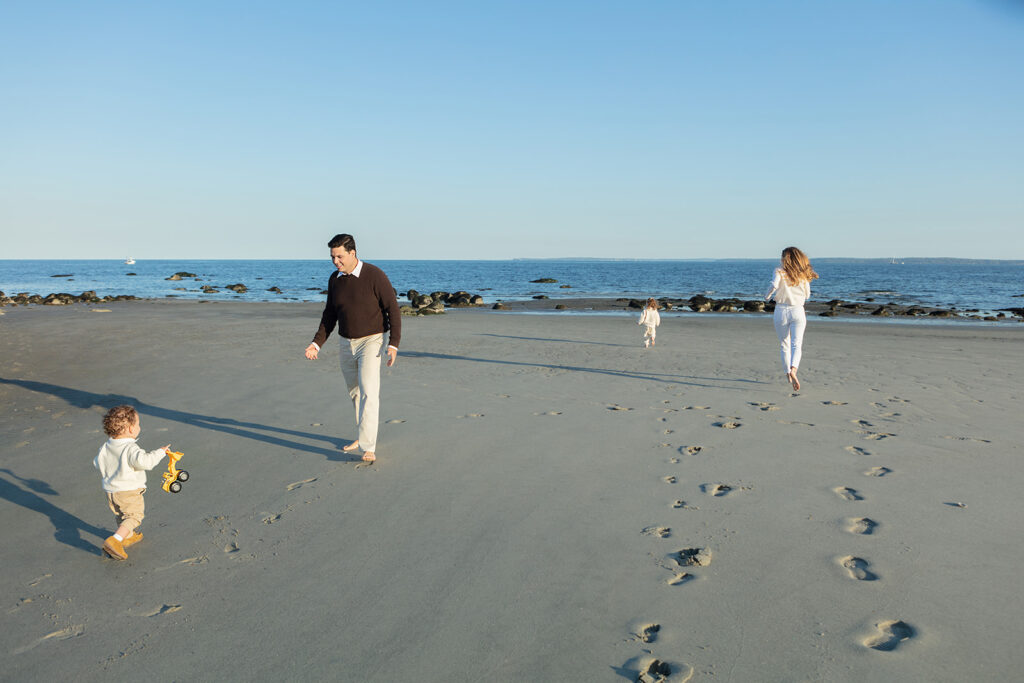 A young family of four creates footprints in the sand along the Connecticut coastline. The toddler brings his dad a yellow digger, as mom and sister run toward the water.