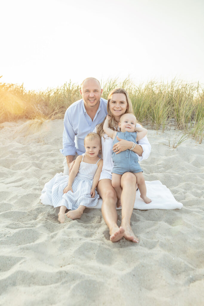 A family of four sit and kneel on a white blanket placed in the soft beach sand of Greenwich Beach, posing for a summer family portrait. The sun sets behind them and natural sun flare leaks into the portrait, giving it a beautiful glow.