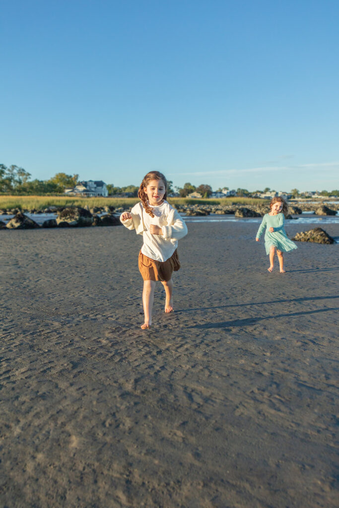 Two little girls run on the dark sandy beach of Tod's Point, coastal rocks, homes, and beach grasses dotting the background. The sky shines bright blue despite the setting sun, and the girls appear as carefree as can be!