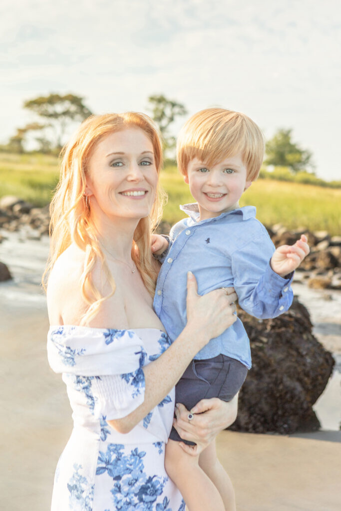A mama with textured, light red hair and gray-blue eyes holds her young son (who looks so much like her!) in her arms. The pair are captured during the Golden Hour on a Connecticut beach.