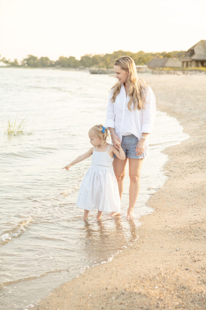 A mother, classically dressed in New England style linen and chambray, walks along Greenwich Beach with her young daughter who wears a seersucker summer dress and a bright blue bow in her hair. 