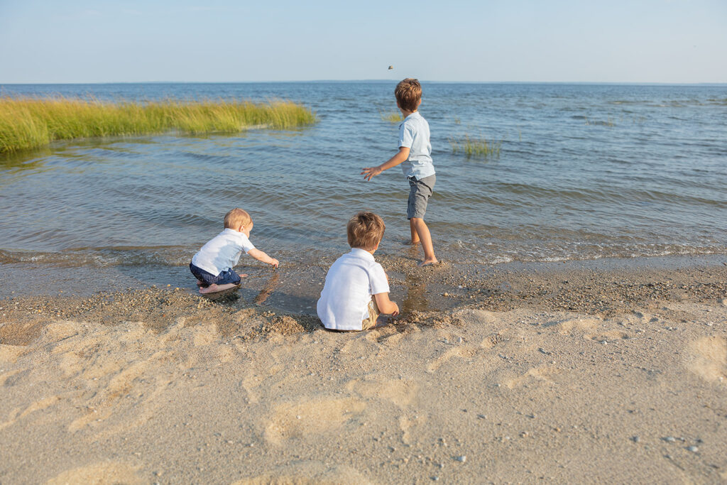 Three young brothers with sandy hair, dressed in classic American outfits of polo shirts with chino shorts, skip rocks into the Atlantic ocean at Greenwich, Connecticut.