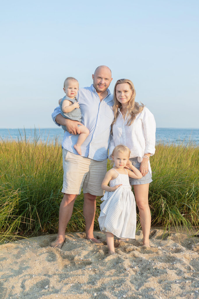 A classic family beach photo. A family of four smile and look at the camera as the setting sun's glow bathes the family in beautiful light. Behind them, beach grasses, the Atlantic ocean, and a clear blue sky.