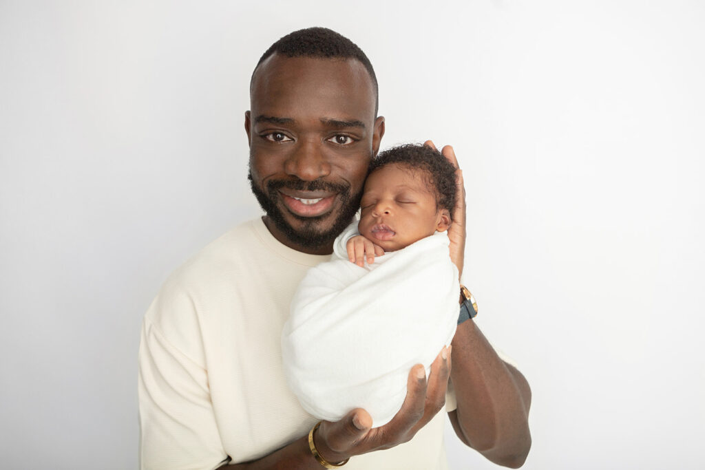 Black father and newborn son portrait. Dad smiles gently and holds his swaddled newborn son in his hands, touching his son's forehead to his cheek.