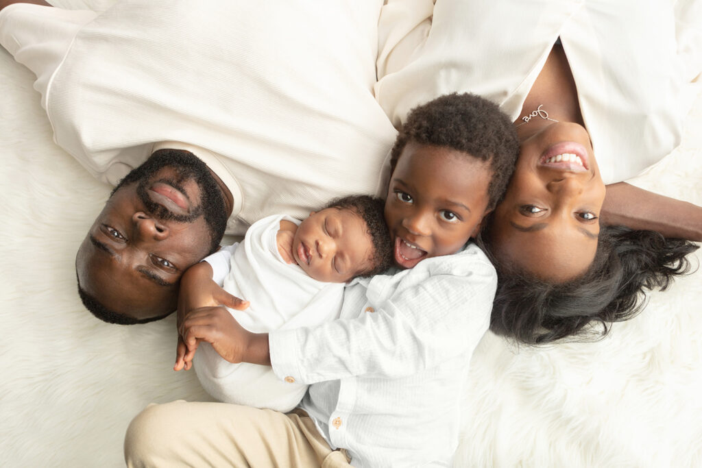 Black family newborn photoshoot. Gorgeous family of four: dad, newborn son, big brother, mom, are photographed looking up from a flokati in the Looking Up Photography studio in Greenwich, Connecticut.
