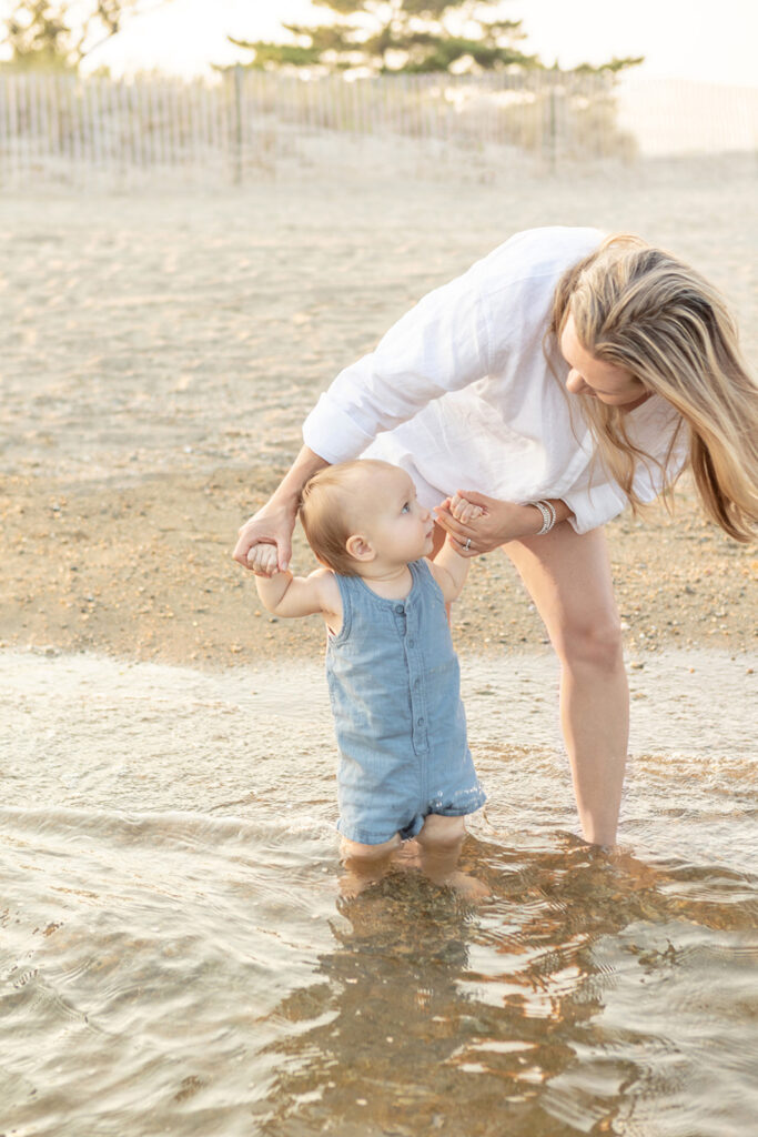 A mother with long blonde hair, wearing a classic white linen shirt, playfully looks down at her baby boy, who looks up at her with a puzzled look on his face. The pair are standing in the shallow water of the Atlantic ocean.
