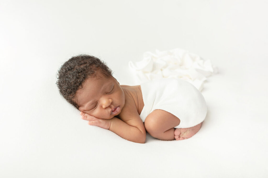 African American newborn baby boy sleeping with his face on his right hand. The baby is lightly swaddled from the waist down in white, the swaddle trailing loosely behind him. 