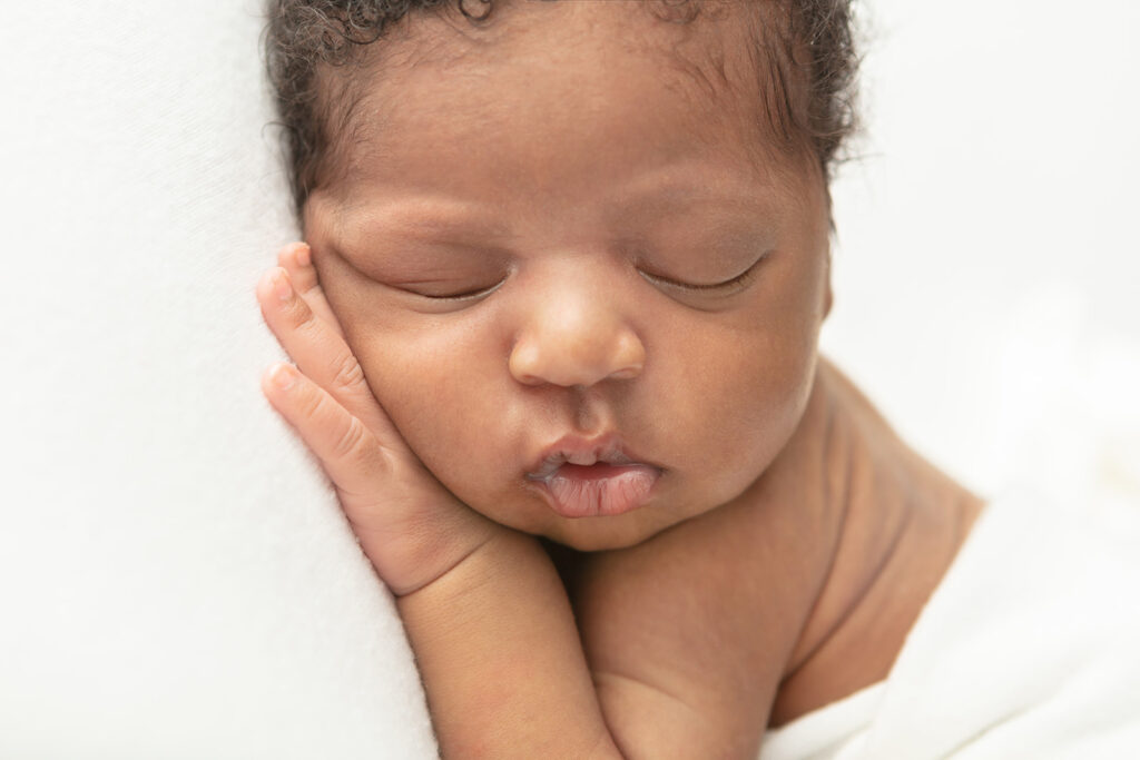 Black baby newborn portrait up close. The baby boy's darling facial features are visible, including curly tendrils around his forehead and ears.