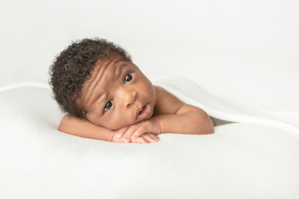 A newborn baby boy with big, dark eyes, rests his head on his folded arms and looks inquisitively at the camera, one eyebrow raised as if to ask the photographer who she is. 