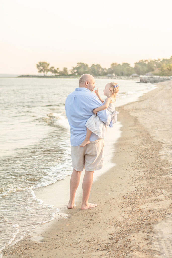 A dad wearing blue oxford and linen shorts walks along the beach, holding his little girl, who wears a seersucker dress tied in the back with a bow, and a blue bow in her hair. The little girl looks as if she's telling her dad something important, holding up her finger in front of his face and looking at him intently.