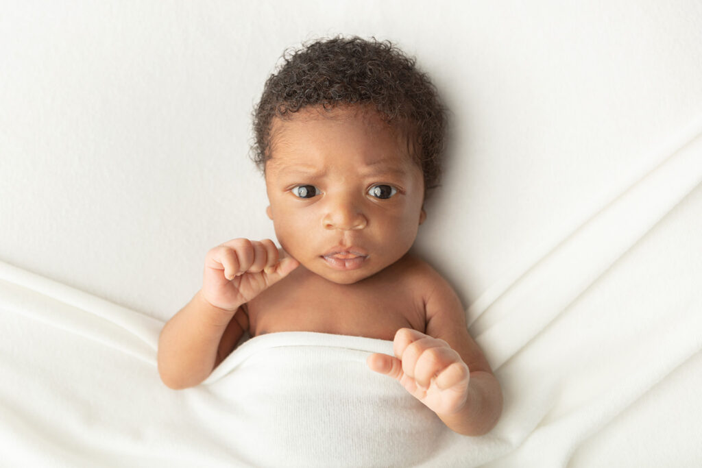 Awake newborn portrait. A baby boy with dark, curly hair is lightly draped in a creamy colored blanket and photographed against an ivory backdrop.