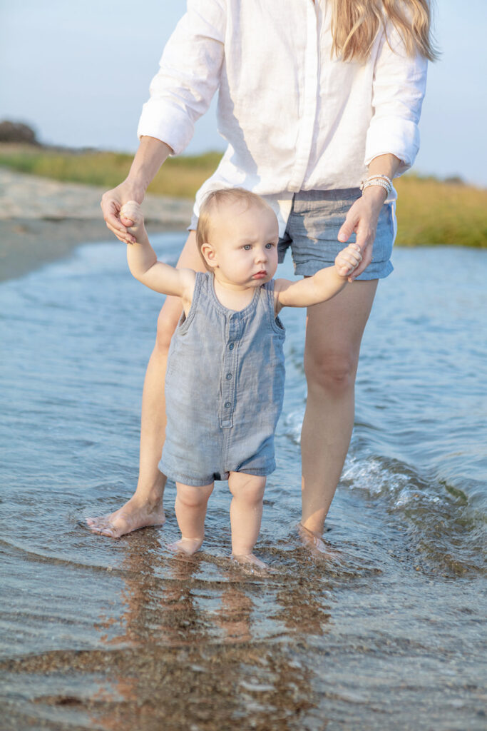 A baby boy wearing a sleeveless, button-up, chambray playsuit, walks seriously along Greenwich Beach. His mom (who's face is out of frame), holds onto his hands, letting him guide her through the shallow water.