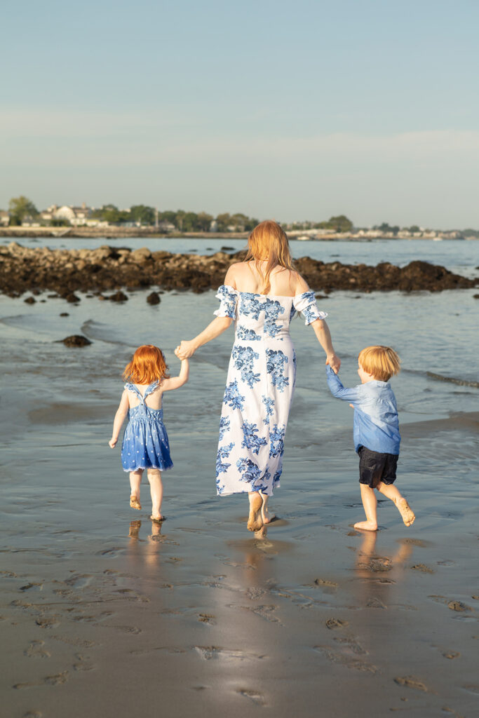 A mother and her two young, red-headed children, walk along a rocky beach in Connecticut. A craggy outcropping is visible in the distance, and the family's footprints are imprinted in the sand.