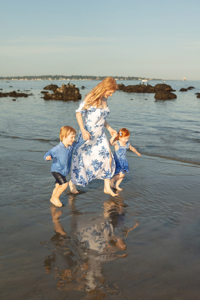 A red-headed trio of mama, her young son and daughter, run happily along a Connecticut beach in the summertime. Behind them, rocks, sea life, and sailboats are visible in the distance.