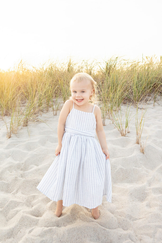 A little girl smiles for a beach portrait, looking as if she is about to twirl in her white and blue seersucker dress. She is framed by wild beach grasses, and a glowing, golden setting sun in the background.