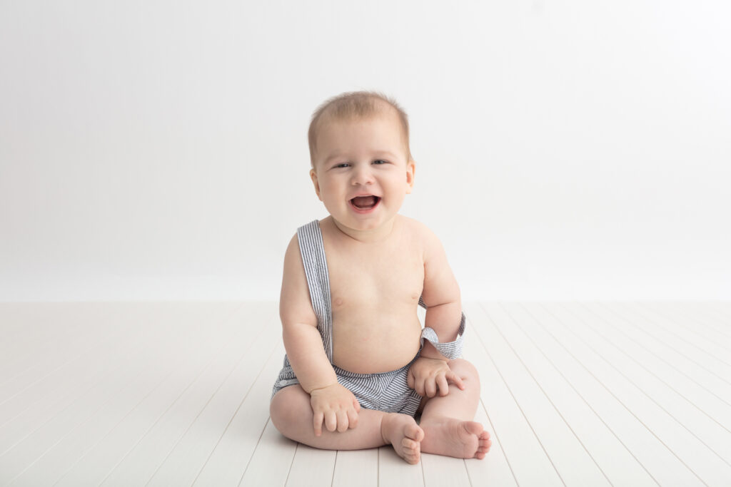A chunky baby sits up on a white wooden plank floor, wearing seersucker suspenders. The baby has fuzzy hair and a big smile on his face.