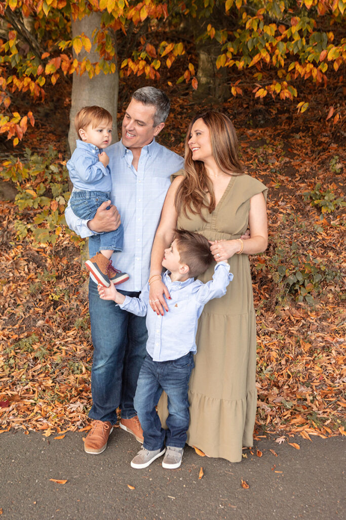 Family of four enjoying a cozy autumn day outdoors in Greenwich, Connecticut, parents embracing their two young sons amidst the golden fall foliage, radiating warmth and love.