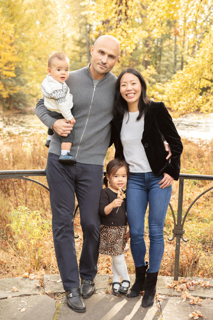Family of four enjoys fall foliage. Mom, dad, toddler girl, and baby amid colorful autumn leaves. Natural beauty and togetherness.