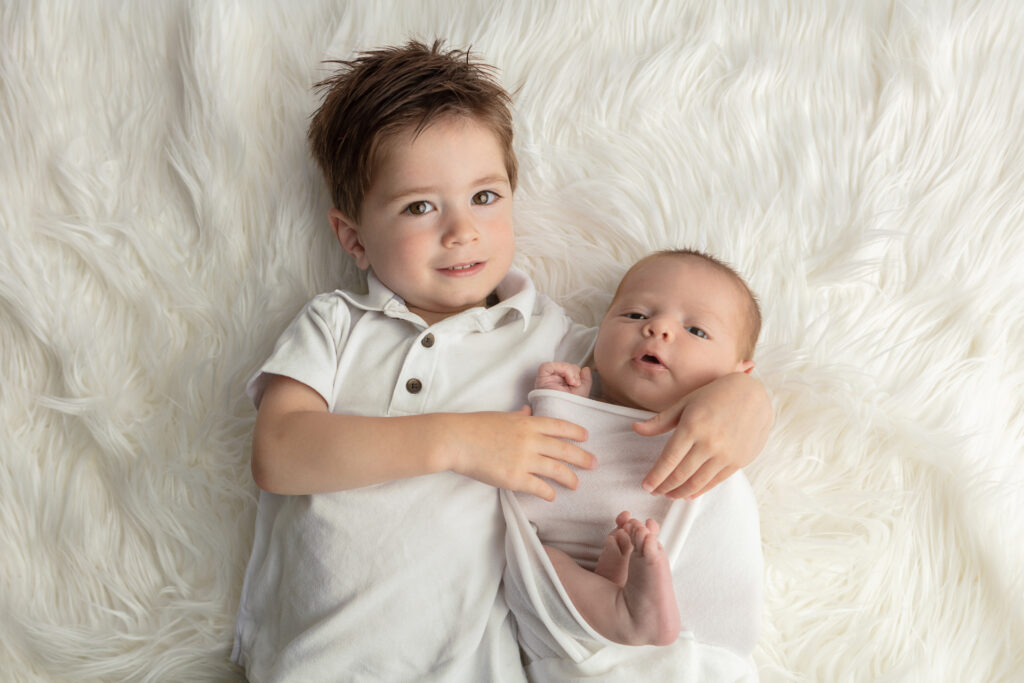 Sibling newborn portrait with two brothers in white. Big brother Austin holds his new baby brother Aiden in his arms as the pair look at the photographer.