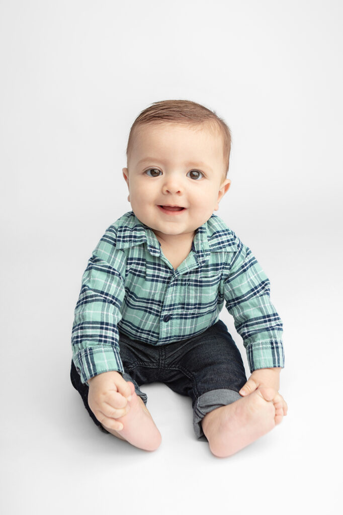 Milestone sitting portrait session. A 6 month old little boy wearing a dusty turquoise flannel shirt and dark, cuffed denim jeans, sits up smiling, holding on to his feet.