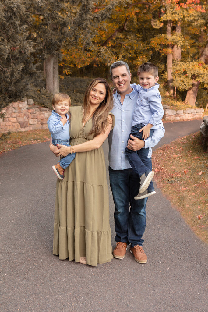 The Deleary family is photographed in a Greenwich, Connecticut park for a fall family portrait session. Mom and dad hold their two young boys in their arms.