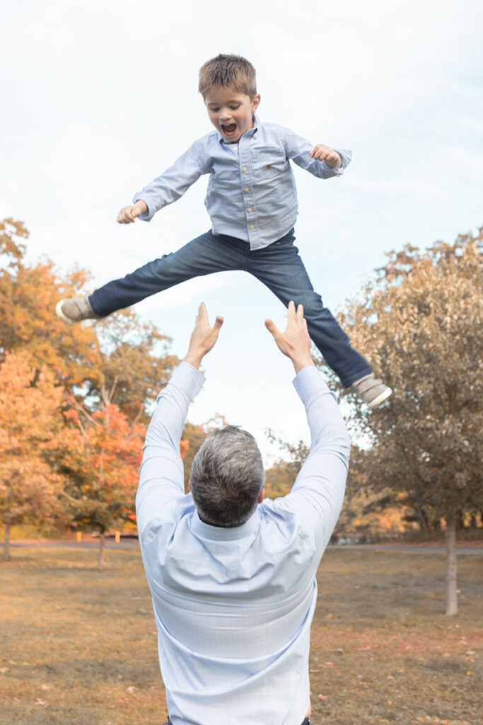 Energetic dad joyfully tossing his giggling young son into the air, capturing a playful and loving father-son moment. Fall family portrait session by Looking Up Photography