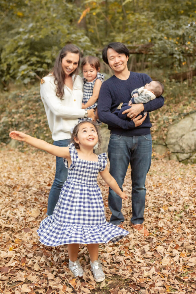 A multicultural family of 5 are photographed in October in beautiful Greenwich, Connecticut. The oldest daughter wears a twirly gingham dress and playfully tosses brown leaves into the air.