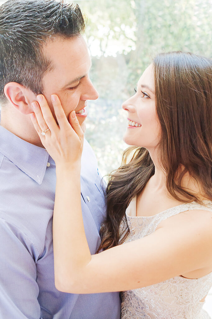 A couple are photographed for their engagement. The newly engaged woman holds her fiance's face with her left hand (showing off her engagement ring) as the duo smile tenderly at each other.