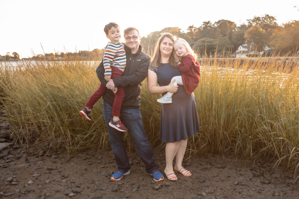 A family of four are photographed during the Golden Hour in the fall in Connecticut. Dad and mom each hold a child in their arms. The family are dressed in shades of navy and maroon.