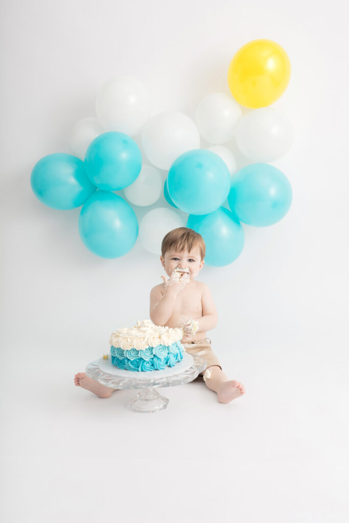 Sunny day inspired milestone cake smash portrait session. A one year-old little boy in khaki shorts tries his first taste of cake frosting, as cloud and sun-inspired balloons hang on the backdrop behind him.