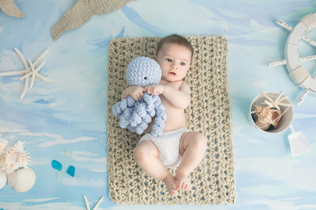Beach inspired 3 month milestone portrait session. The baby boy lies on a jute-looking mat and holds a crocheted octopus in his arms as he looks slightly off-camera.