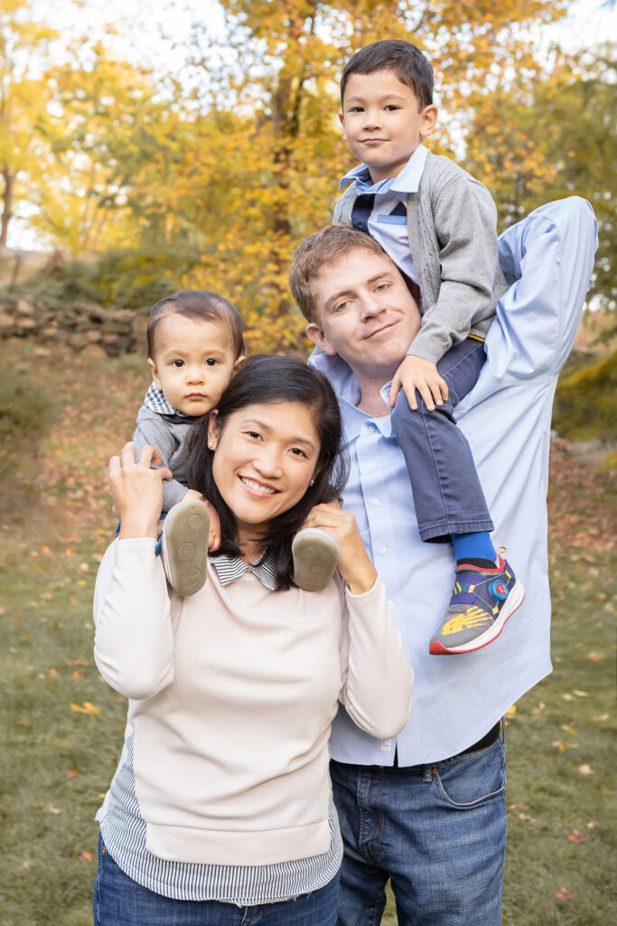 A classically dressed family of four are photographed in a park in Greenwich, Connecticut. Both parents hold their young boys up on their shoulders for the portrait.