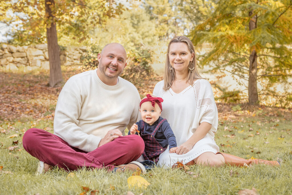 A family of three are photographed for their fall foliage family session by Karen Kahn of Looking Up Photography. Mom and dad are dressed in neutrals while their baby girl brings a pop of fall color to the portrait.
