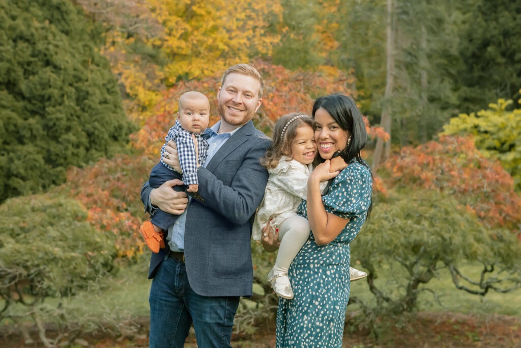 Multicultural family of four enjoys fall foliage. Smiling mom, dad, baby, and toddler embrace amid colorful autumn leaves. 