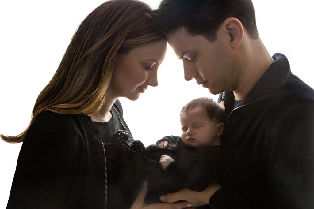 A new mom and dad hold their newborn daughter in between them. The trio are dressed in black and paint a peaceful picture of a growing family.