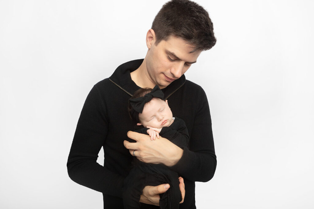A new dad wearing a black, mock-neck zip sweater, holds his newborn baby daughter in his hands and looks down at her peacefully. The newborn baby girl is also wearing black and wears a black bow on her head.