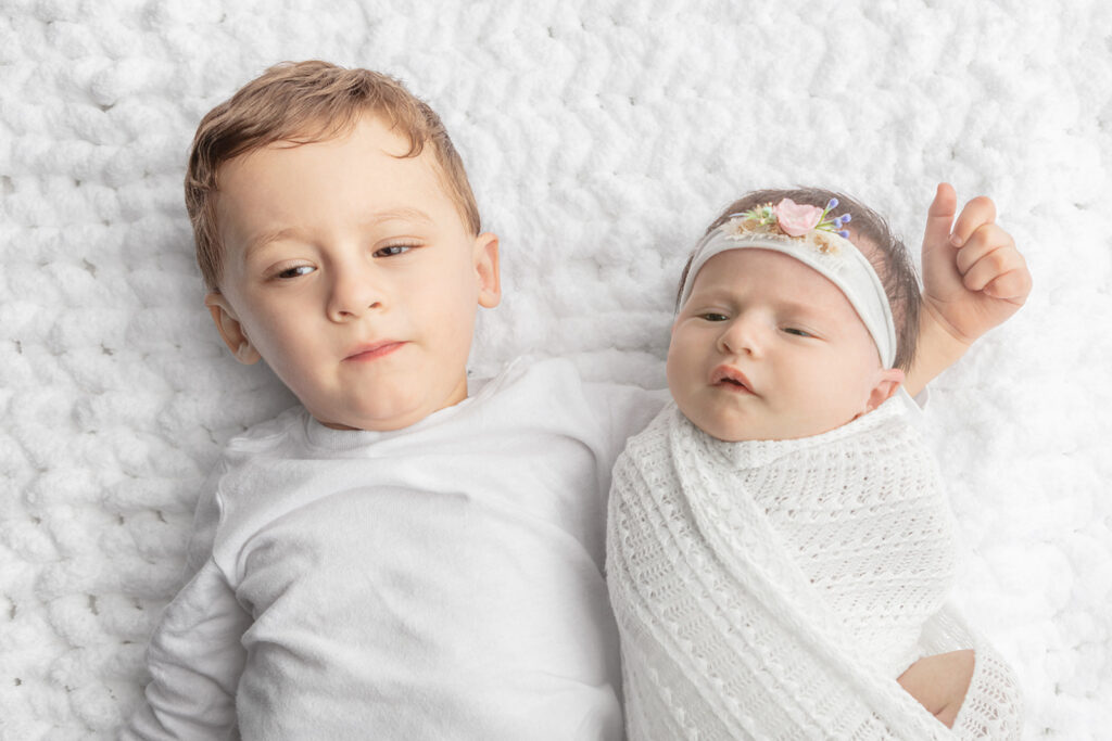 A toddler brother in a long sleeved white t-shirt holds his arm around his newborn baby sister, who is swaddled in a crochet, white blanket. The pair lie on a textured white blanket in the Looking Up Photography studio.