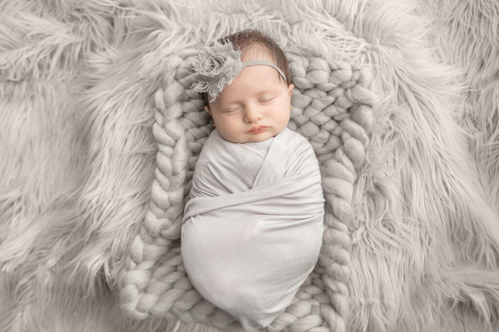 A newborn baby girl sleeps peacefully on layers and different textures of soft gray textiles: flokati, large knit pillow, and finally, her stretchy swaddle blanket.