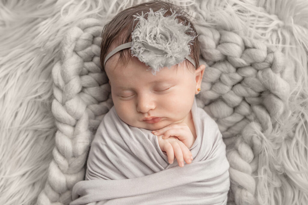 A newborn baby girl with her ears pierced sleeps peacefully with her folded hands peeking out of her gray swaddle blanket. She wears a shabby chic, gray rose headband on her head.