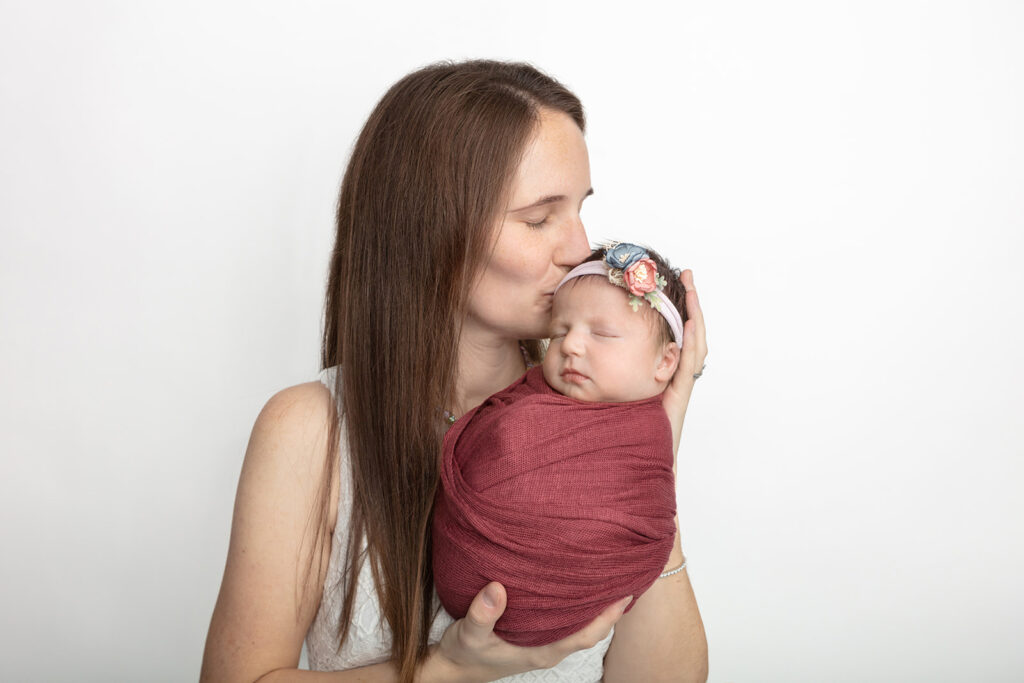 A mother holds up her newborn baby daughter to give her a kiss on the head. Her daughter is wrapped in a dusty burgundy swaddle and wears a floral headband