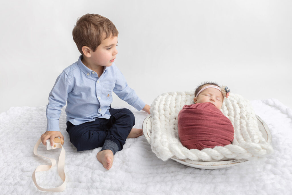 Siblings are photographed in the Looking Up Photography studio. Big brother wears a classic light chambray shirt and dark denim pants as he looks down at his new baby sister.