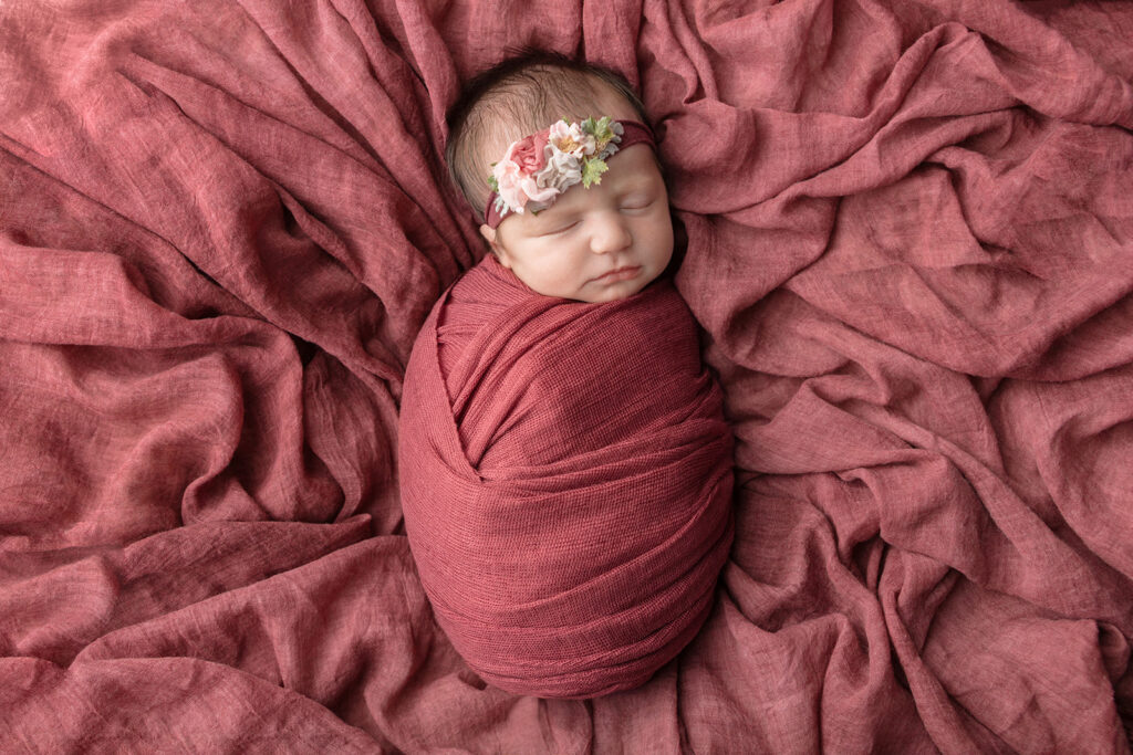A newborn baby girl, tightly swaddled in dusty burgundy, is photographed wearing a stretchy floral headband, on a rumpled dusty rose linen sheet