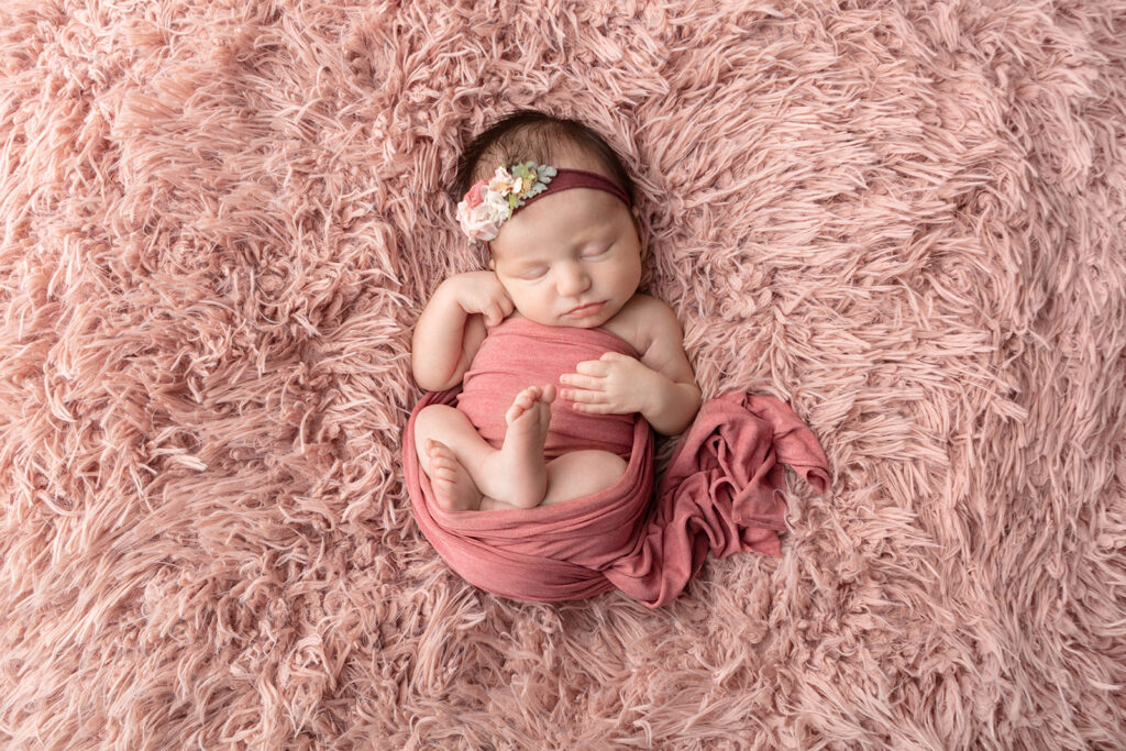 Newborn baby girl lightly swaddled in dusty rose, photographed on a dusty pink flokati. Photographed in the Looking Up Photography studio in Greenwich, Connecticut