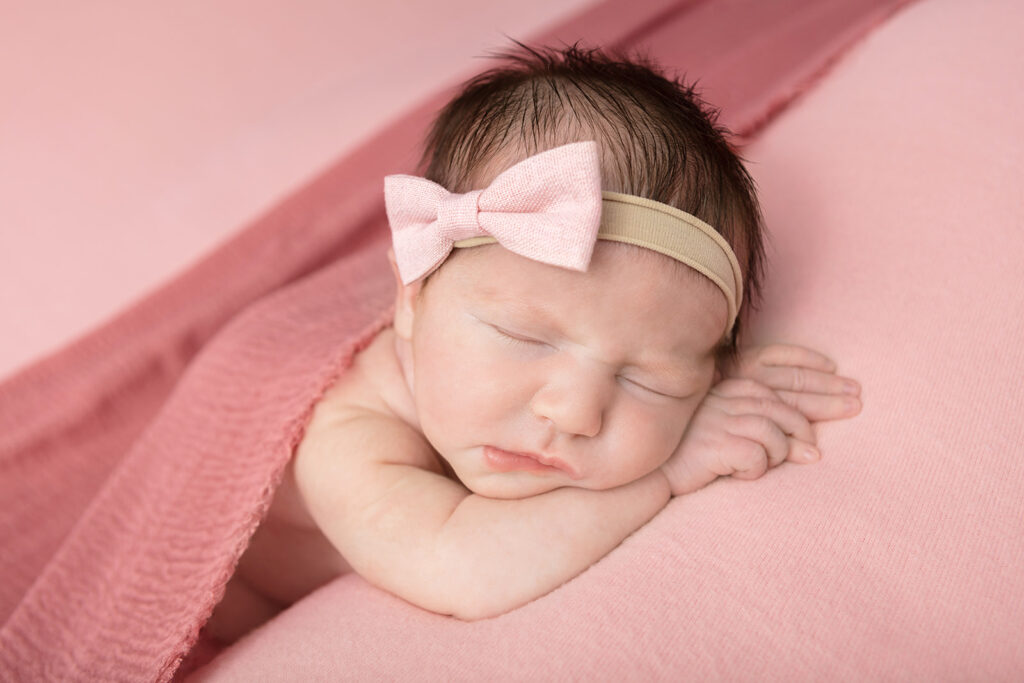 A newborn baby girl, Natalie, is photographed on soft, light pink fabric, with her hands folded underneath the side of her face. She wears a light pink bow headband