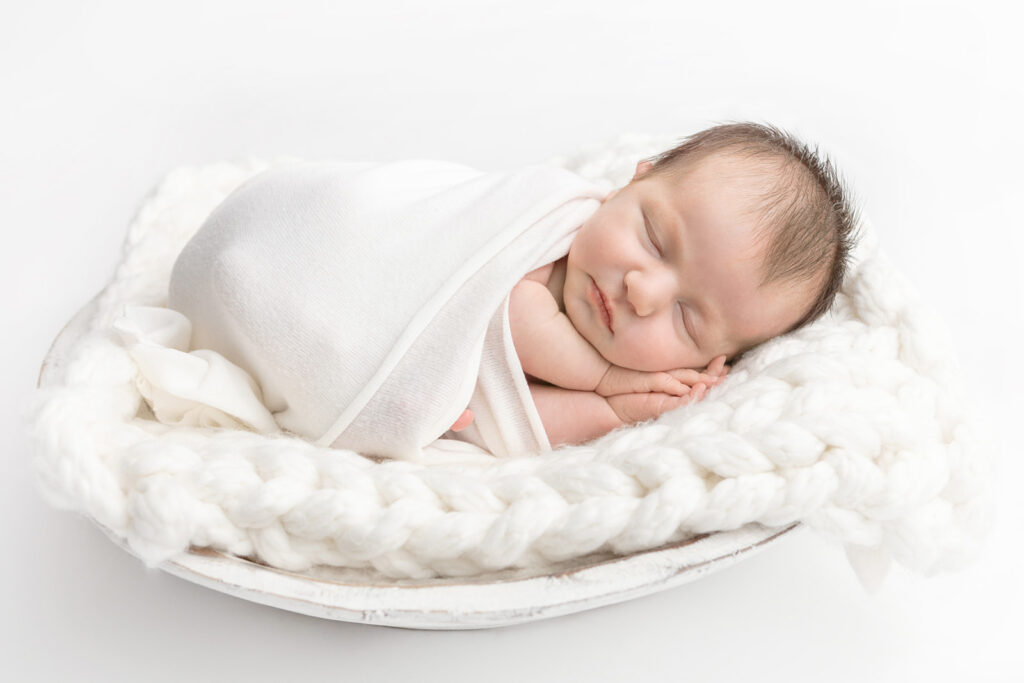 A newborn baby girl, swaddled in soft ivory, is photographed on a chunky knit blanket square in a distressed milk bowl. Photographed by Karen Kahn in the Looking Up Photography studio